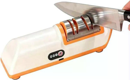 An electric knife sharpener with a white and orange plastic body, shown sharpening a stainless steel kitchen knife. A human hand is visible holding the knife, which classifies the subject type as Lifestyle.